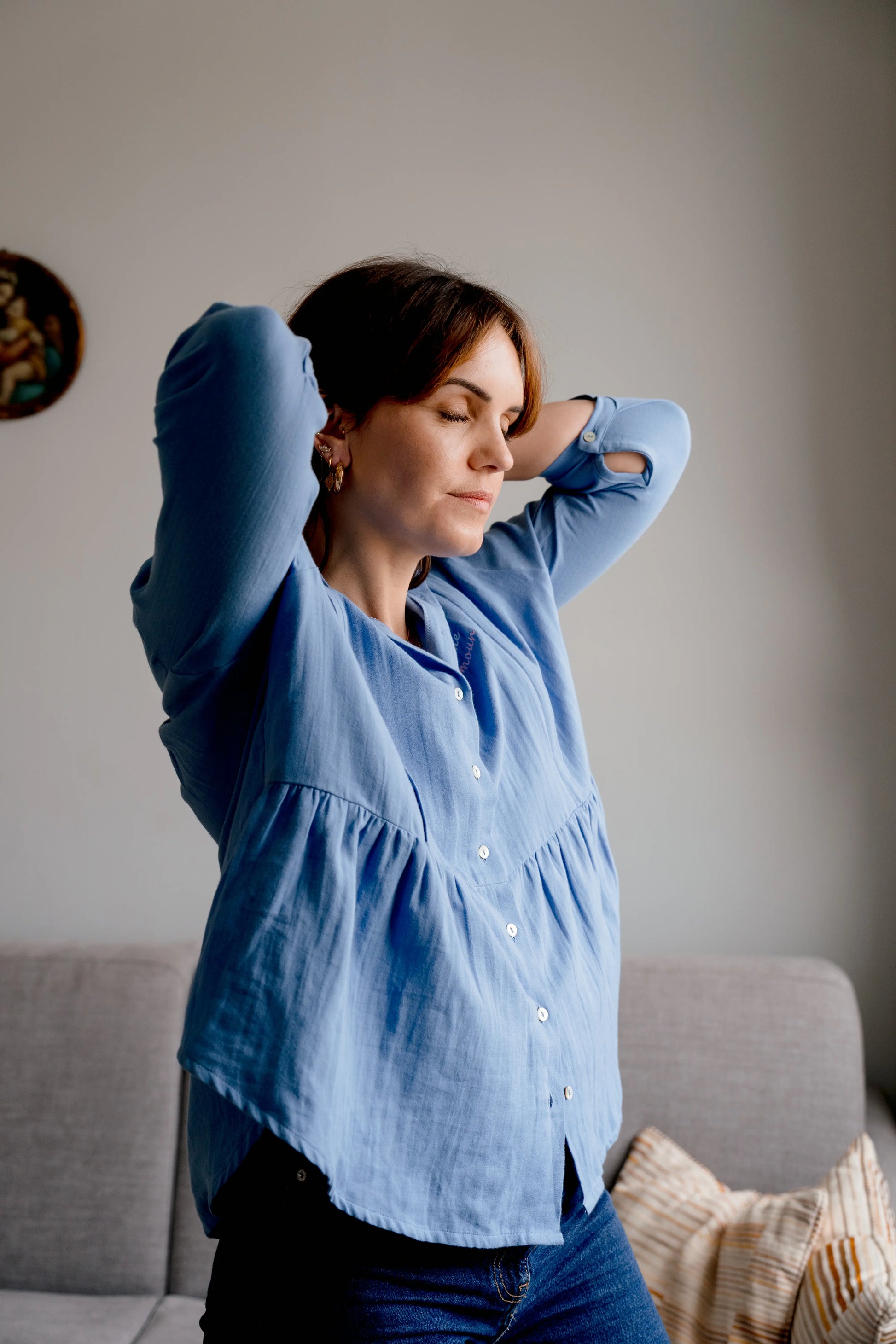 Blouse bleu de grossesse et allaitement pour vivre pleinement sa maternité et nourrir son bébé à la demande. La maman allaite son bébé confortablement dans un fauteuil chez elle, et caline son bébé.