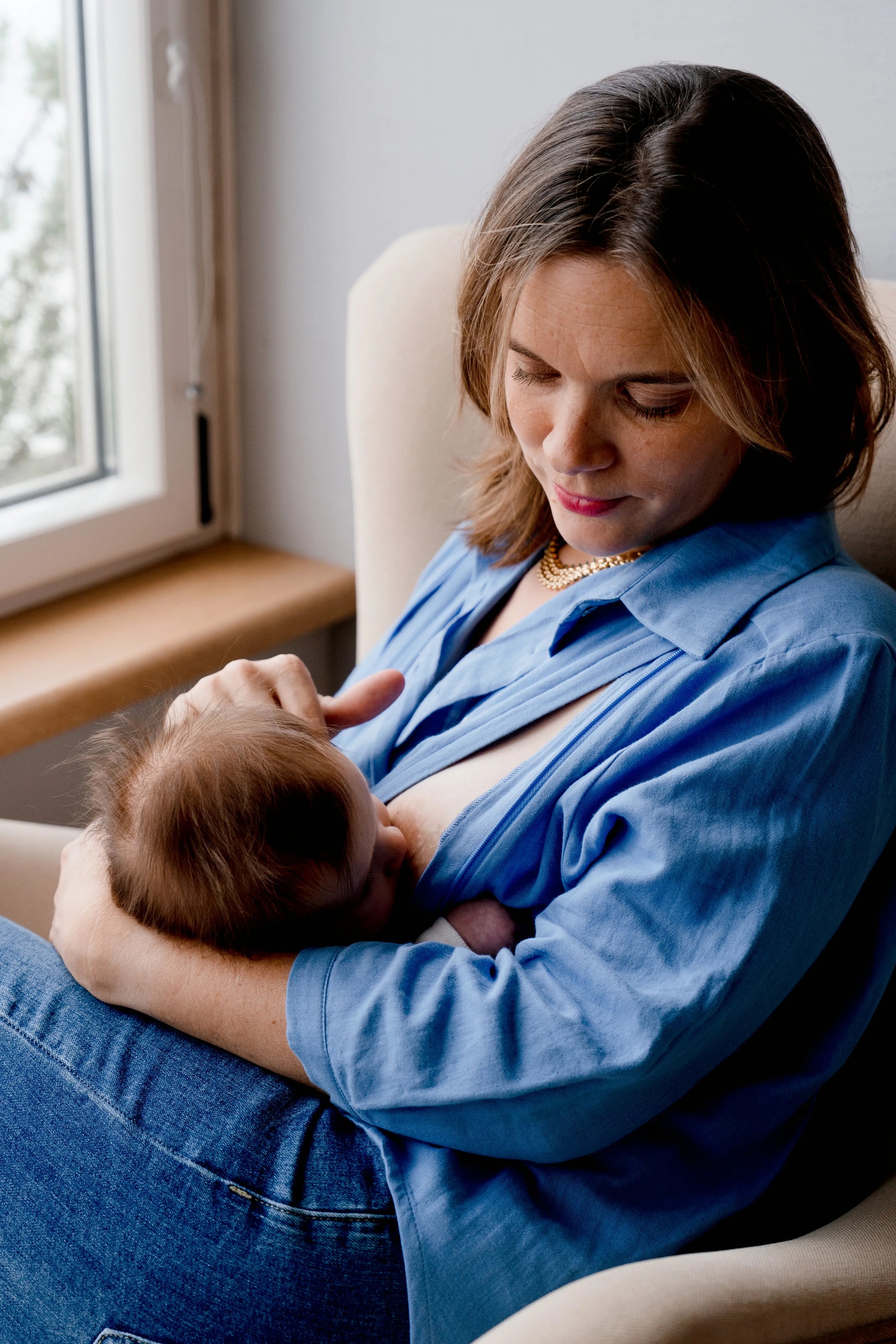 Blouse de grossesse et allaitement pour vivre pleinement sa maternité et nourrir son bébé à la demande. La maman allaite son bébé confortablement dans un fauteuil chez elle, et caline son bébé.