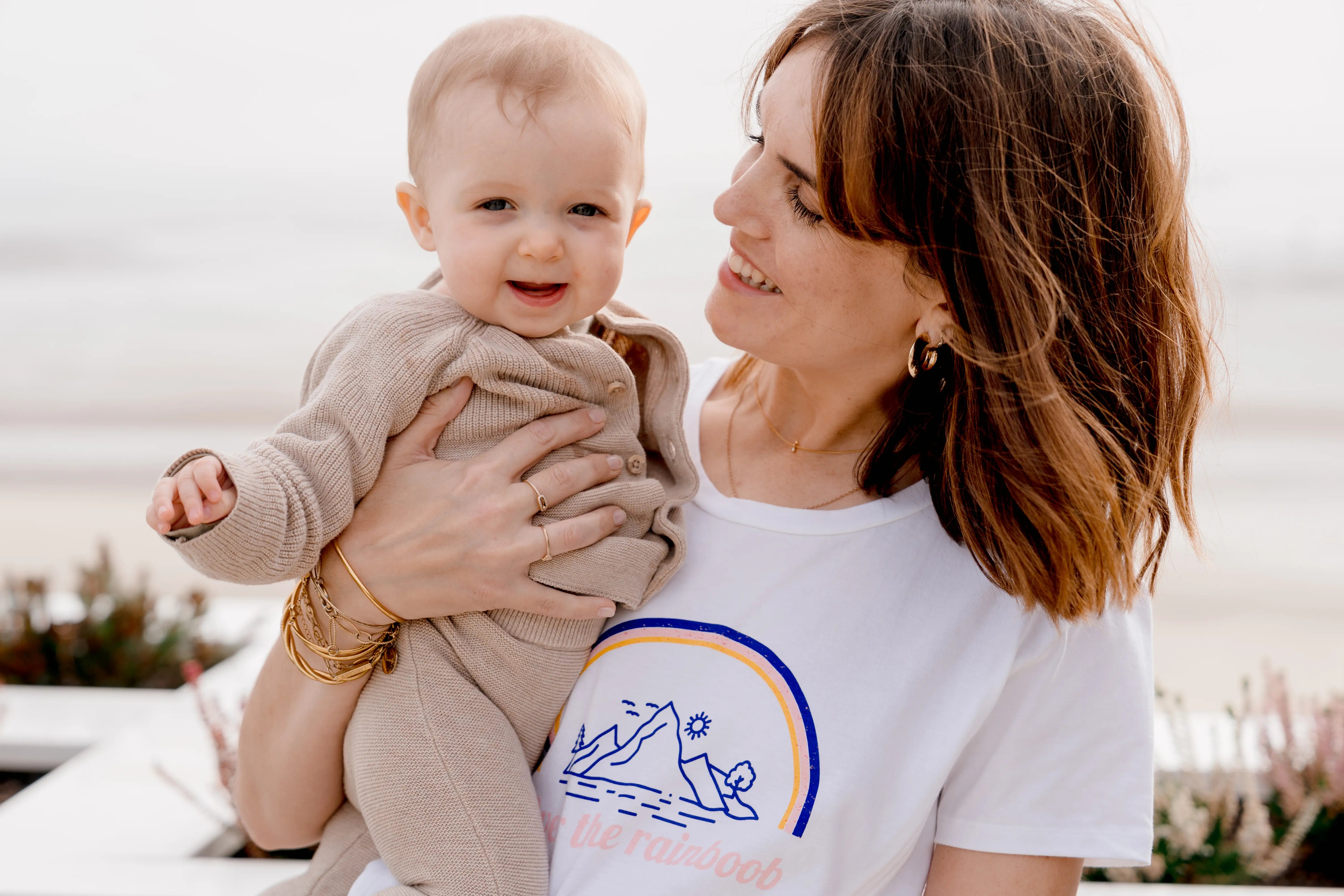 Maman qui porte son bébé et qui lui sourit. Elle porte un T-shirt allaitement 100% coton, avec des ouvertures avec zips pour allaiter facilement et discrètement. design 